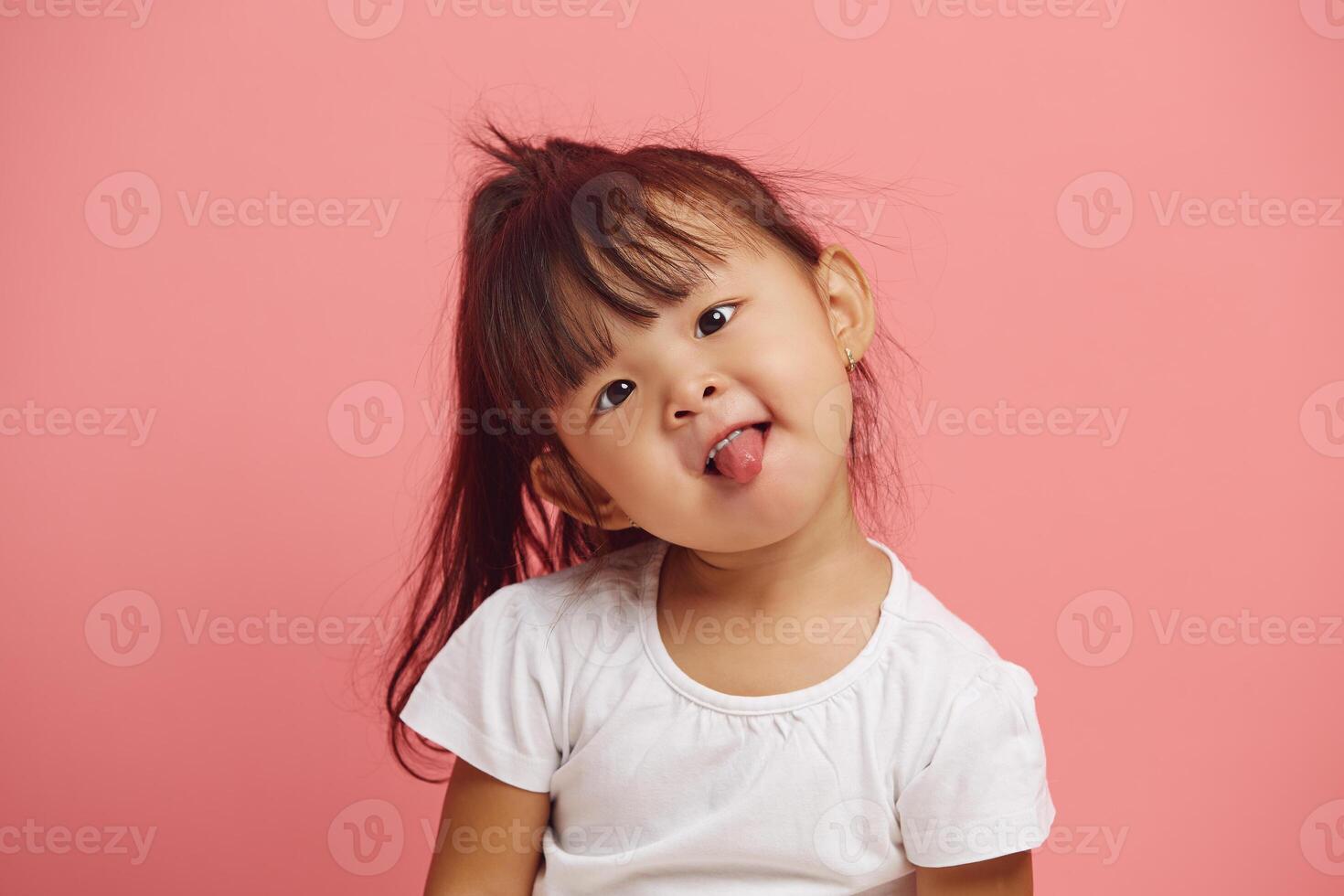 headshot portrait of playful female child tilted her head and shows her tongue standing over a pink isolated background. photo