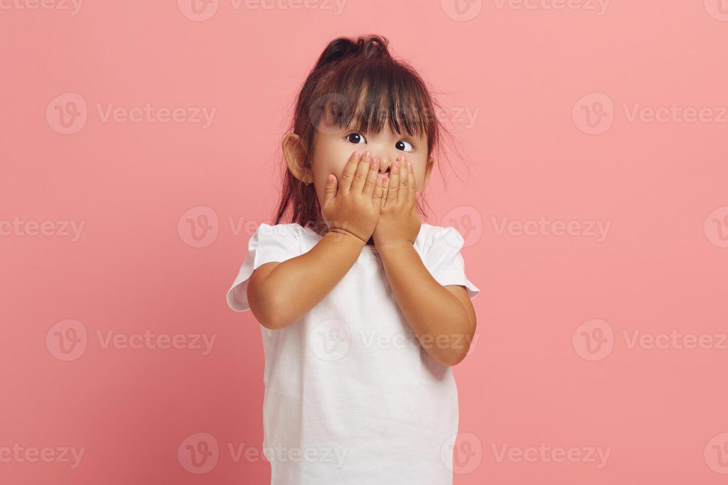 Shocked little girl covers mouth with hands stares scared at camera cannot believe in something horrible dressed in white t shirt isolated over pink background. photo