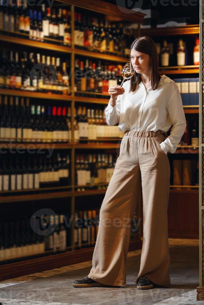 Experienced female sommelier smells wine into glass standing in cellar over plenty bottle shelf background. photo