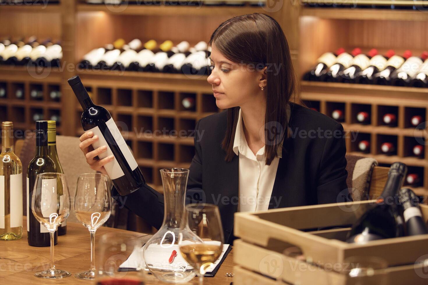 Focused female wine expert during tasting wearing in classic clothes holding bottle of red wine and reads label with characteristics of wine, the year of manufacture of the alcoholic beverage. photo