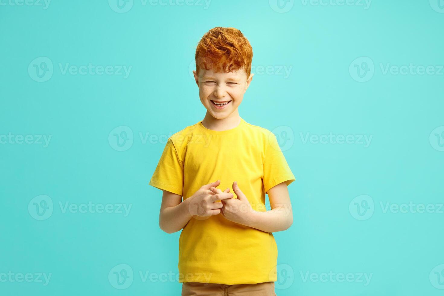 Irish Red-Haired Boy in Yellow T Shirt Laughing and Making a Playfully Shyness Gesture Against Bright Blue Isolated Background. photo
