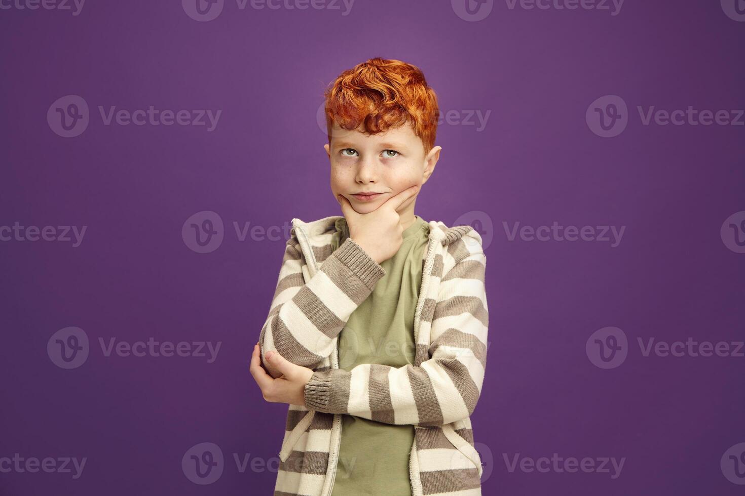 Pensive Red Headed Little Boy Around 7-8 years Puzzled Standing Keeping Hand to Cheek with Thoughtful Face Expression Looking Upwards, Wearing in Casual Clothes Standing on Isolated Violet Background photo