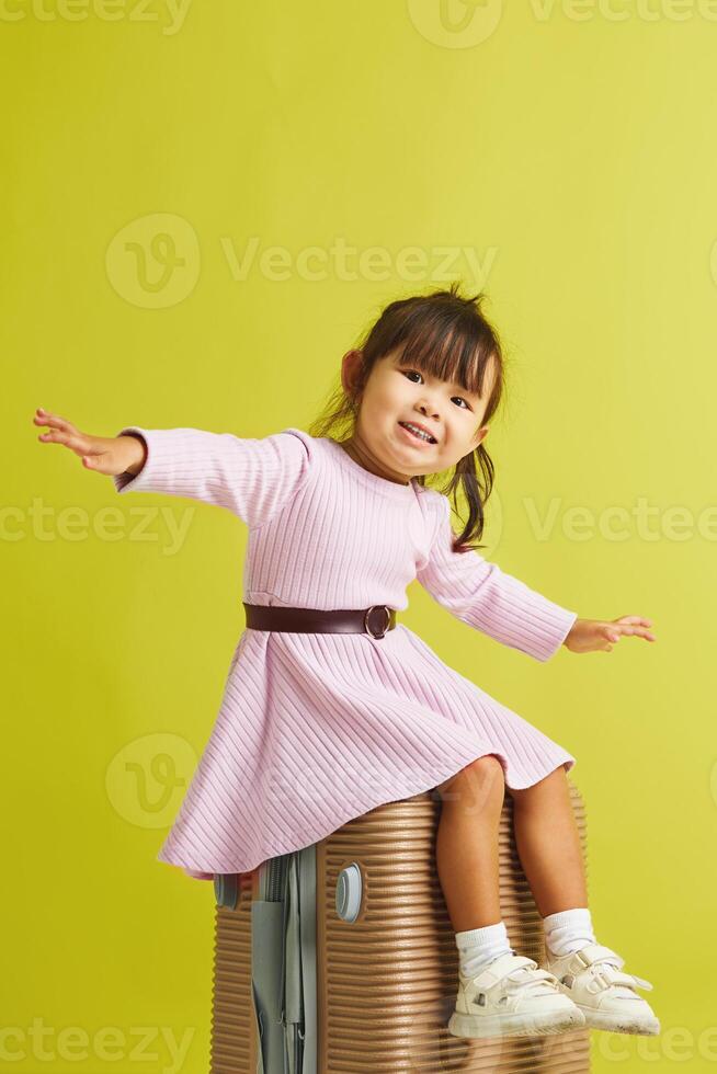 Small asian girl sitting on suitcase and expresses happy anticipation before starting vacation, cheerful waving his arms to the sides, imitating the flight of an airplane over yellow isolated. photo