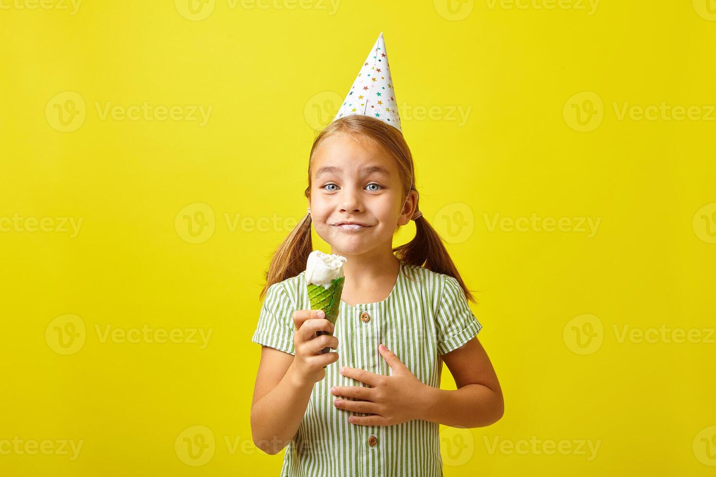 Joyful little girl in a birthday hat, has a surprised expression, eating ice cream, stands on a yellow isolated background. photo