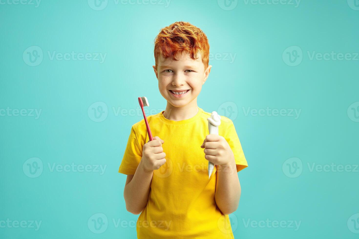 Happy red headed little Irish ethnicity boy holding squeezed toothpaste tube and toothbrush, joyful smiling, wearing in yellow t shirt, standing over blue isolated background. photo