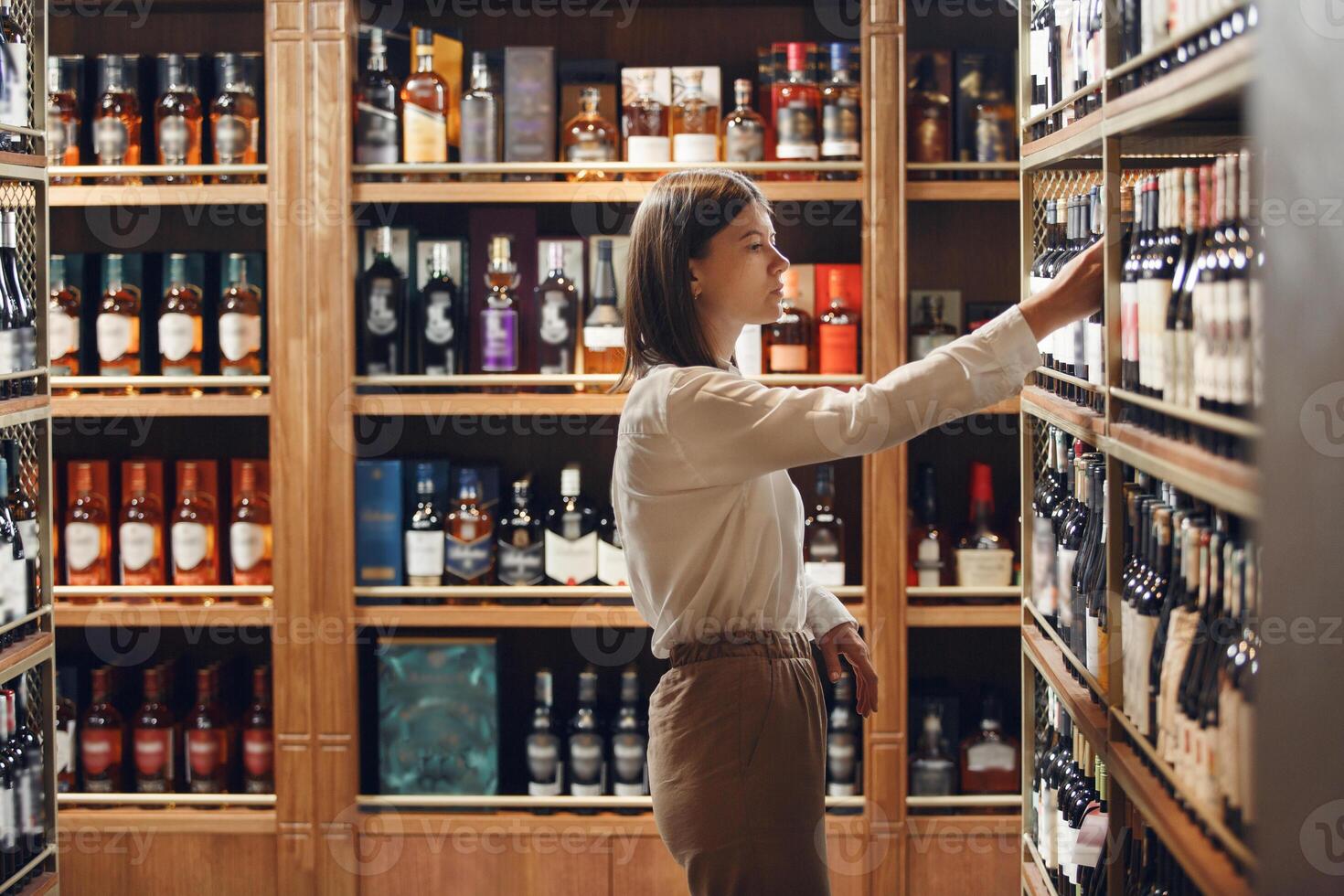 Professional female sommelier choosing wine being at wine cellar full of bottles with exquisite alcohol drinks, that have various sweet and sour tastes on large wooden shelves. photo