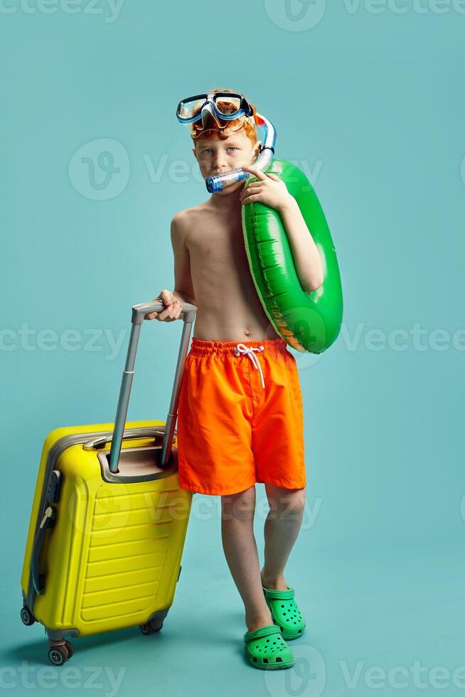 Vertical Shot of Red Headed Child Boy in Orange Swimming Trunks, with Inflatable Ring Around Himself, Having Underwater Mask and Snorkel Holding Travel Suitcase Standing on Blue Isolated Background. photo