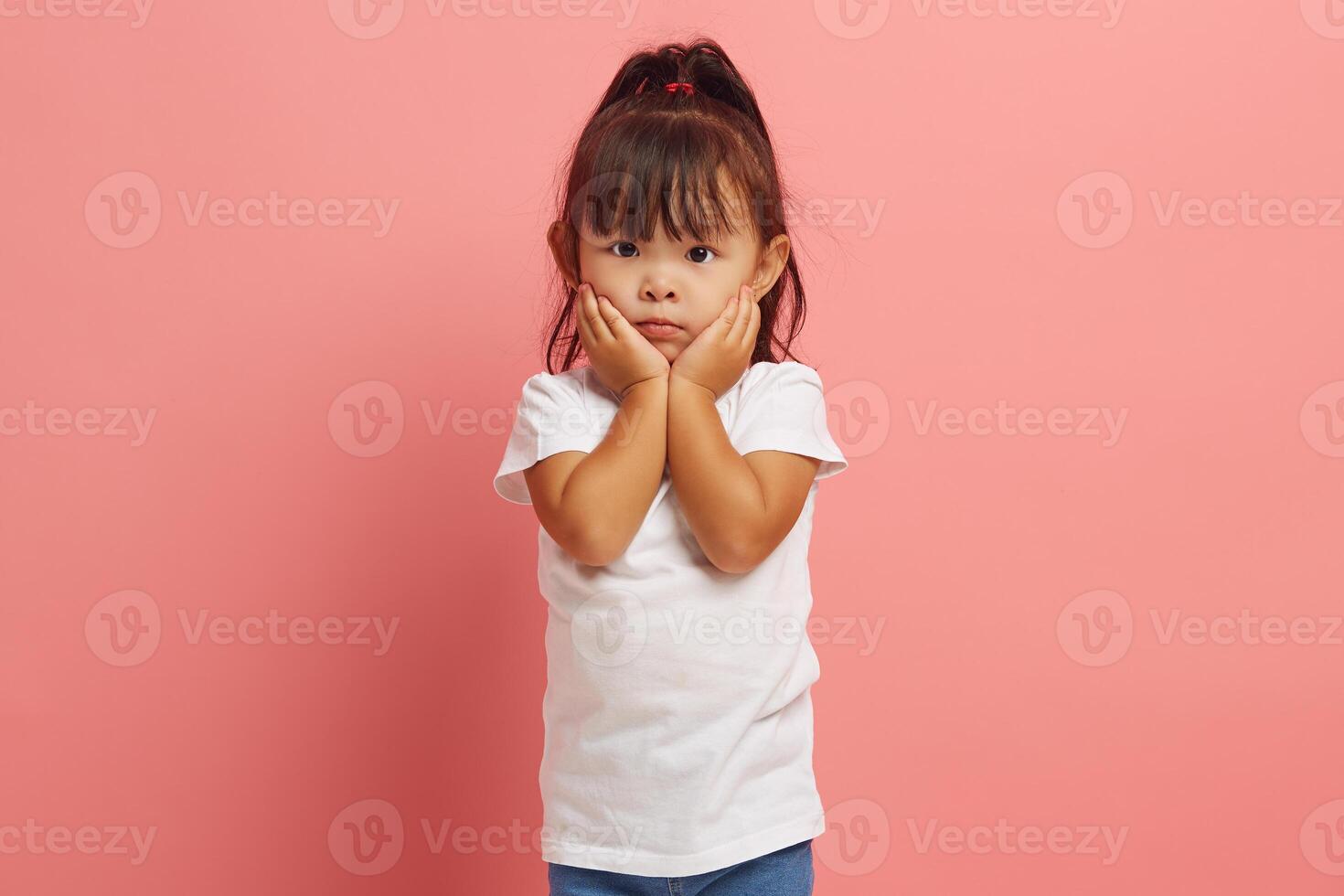 Scared little Asian ethnicity girl looks with fear eyes at camera, being afraid of something, keeps hands on cheeks, looks in terror, isolated over pink background. photo