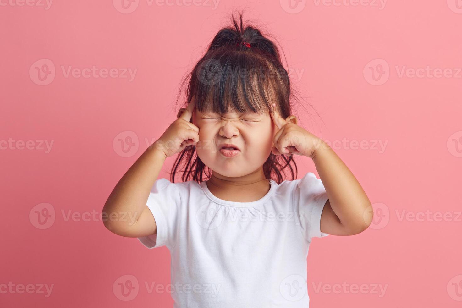 Serious little asian ethnicity girl keeps fingers on temples and closes eyes to remember something important standing isolated over pink background. photo