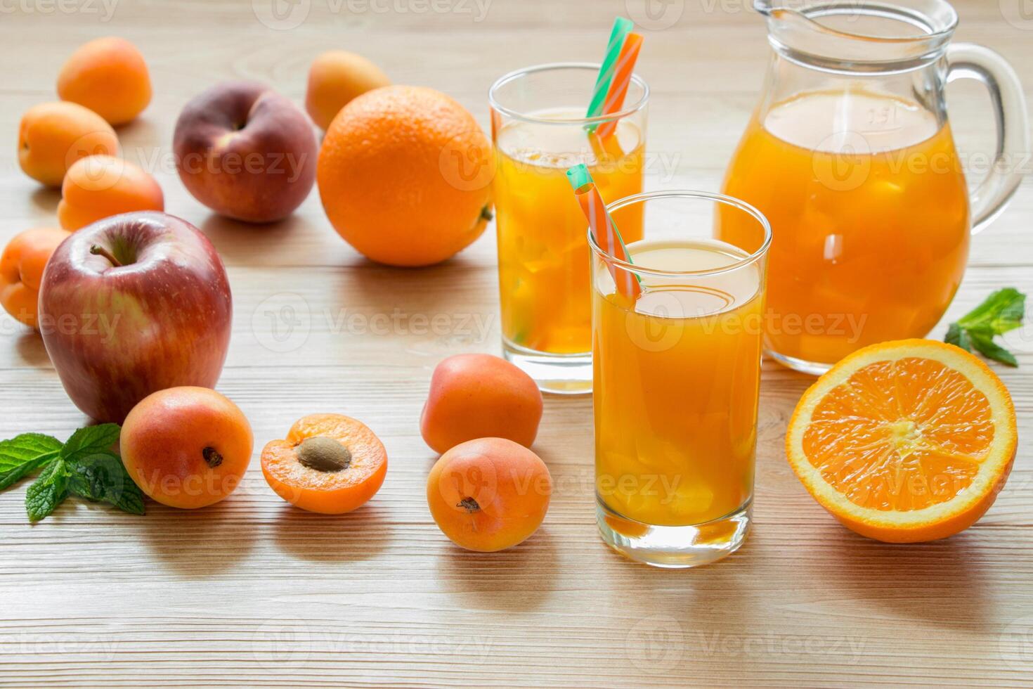 Right fruit juice in glasses with ice near the jar of juice and scattered apricots, peaches, apple, orange left empty space on light wooden background. Apricot peach apple orange juice with ice. photo