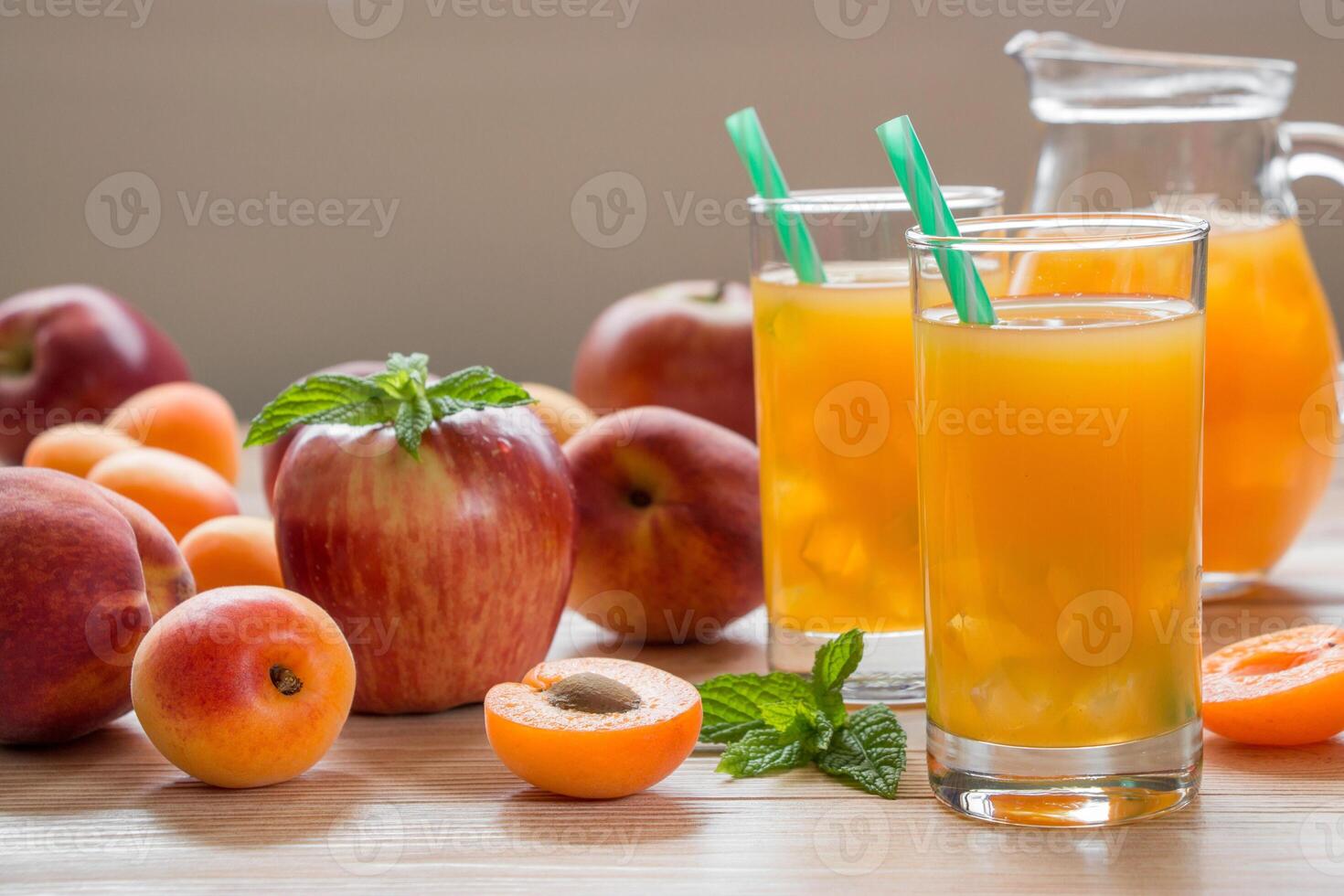 Apricot peach apple juice in glasses with ice near jar of juice and scattered apricots, peaches, apple, mint on a light wooden background. Apricot peach apple juice with ice. Horizontal. Daylight. photo