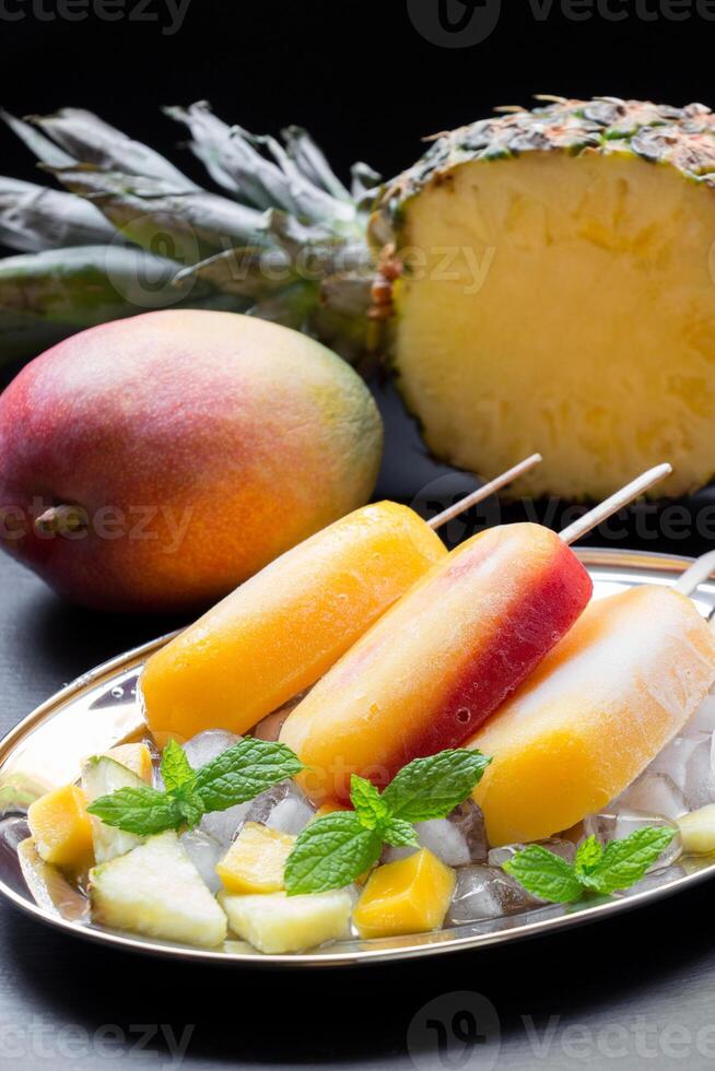 Mango pineapple ice cream popsicles arranged on a decorative plate with crushed ice and fresh fruit. A ripe mango and a halved pineapple enhance the tropical theme. photo