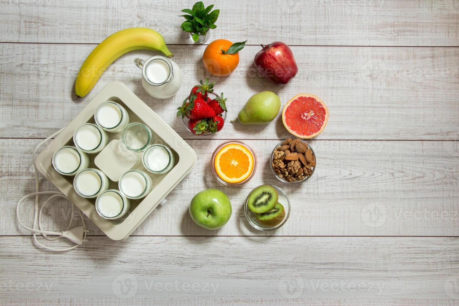 A vibrant collection of fruits including bananas, apples, and berries is placed on a wooden surface alongside a yogurt maker, promoting healthy eating and preparation. photo