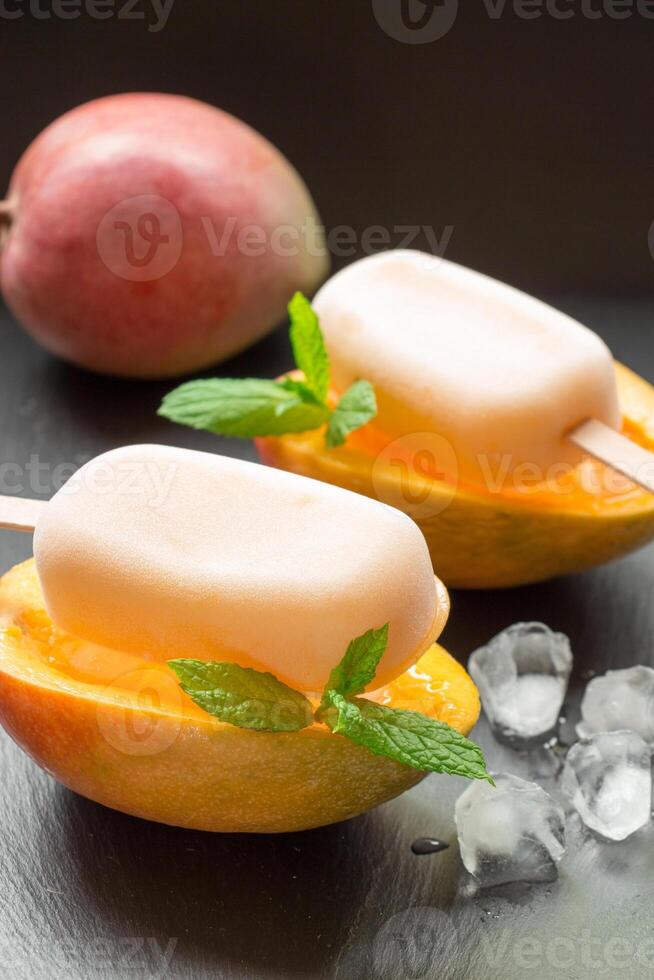 Mango sorbet ice cream on half cutted mango fruit behind mango near ice cubes on a black background. Mango fruit sorbet ice cream popsicles. Vertical. photo