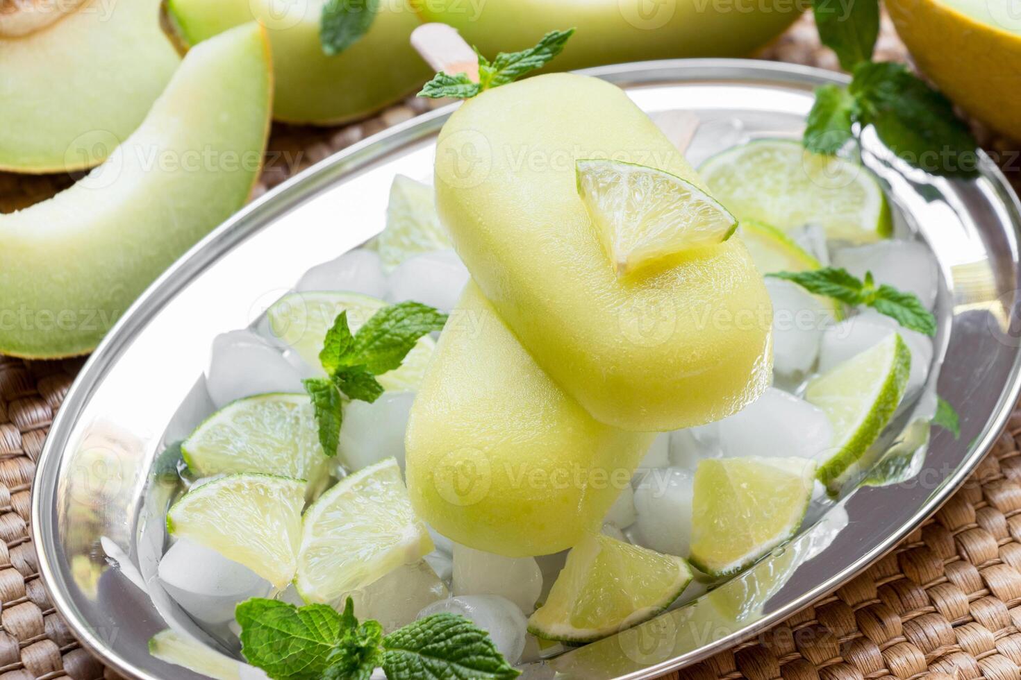 Melon and lime sorbet ice cream on silver plate with ice cubes, lime pieces, mint leaves near melon slices, lime on a straw napkin background. Melon and lime sorbet ice cream popsicles. Horizontal. photo