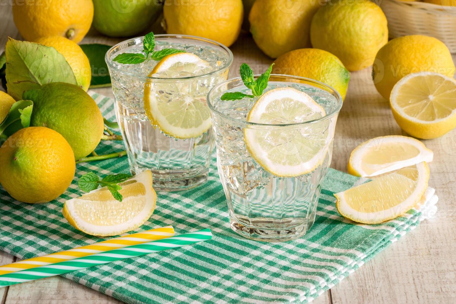 Two glasses filled with lemon drinks sit on a green checkered cloth surrounded by fresh lemons and mint leaves in a sunny kitchen atmosphere, perfect for summer refreshment. photo