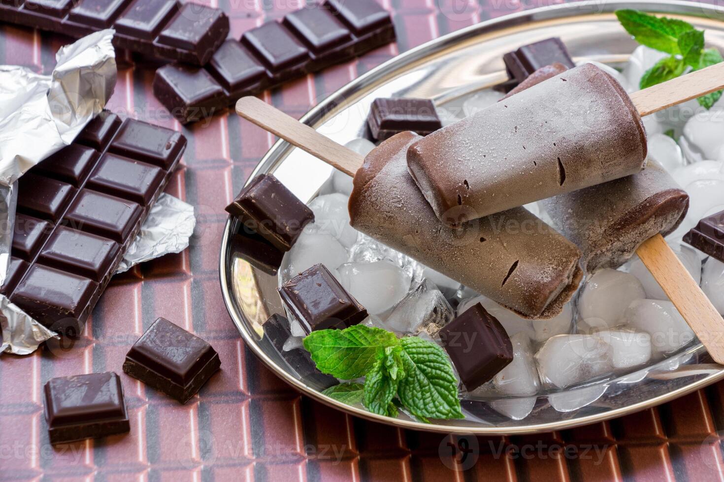 Chocolate ice cream on a stick on a silver platter with ice and pieces of chocolate mint leaves near pieces of chocolate on a chocolate background. Chocolate ice cream popsicles. Horizontal. Close. photo