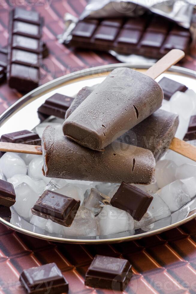 Chocolate ice cream on a stick on a silver platter with ice cubes and pieces of chocolate near pieces of chocolate on a chocolate background. Chocolate ice cream popsicles. Vertical. Close. photo