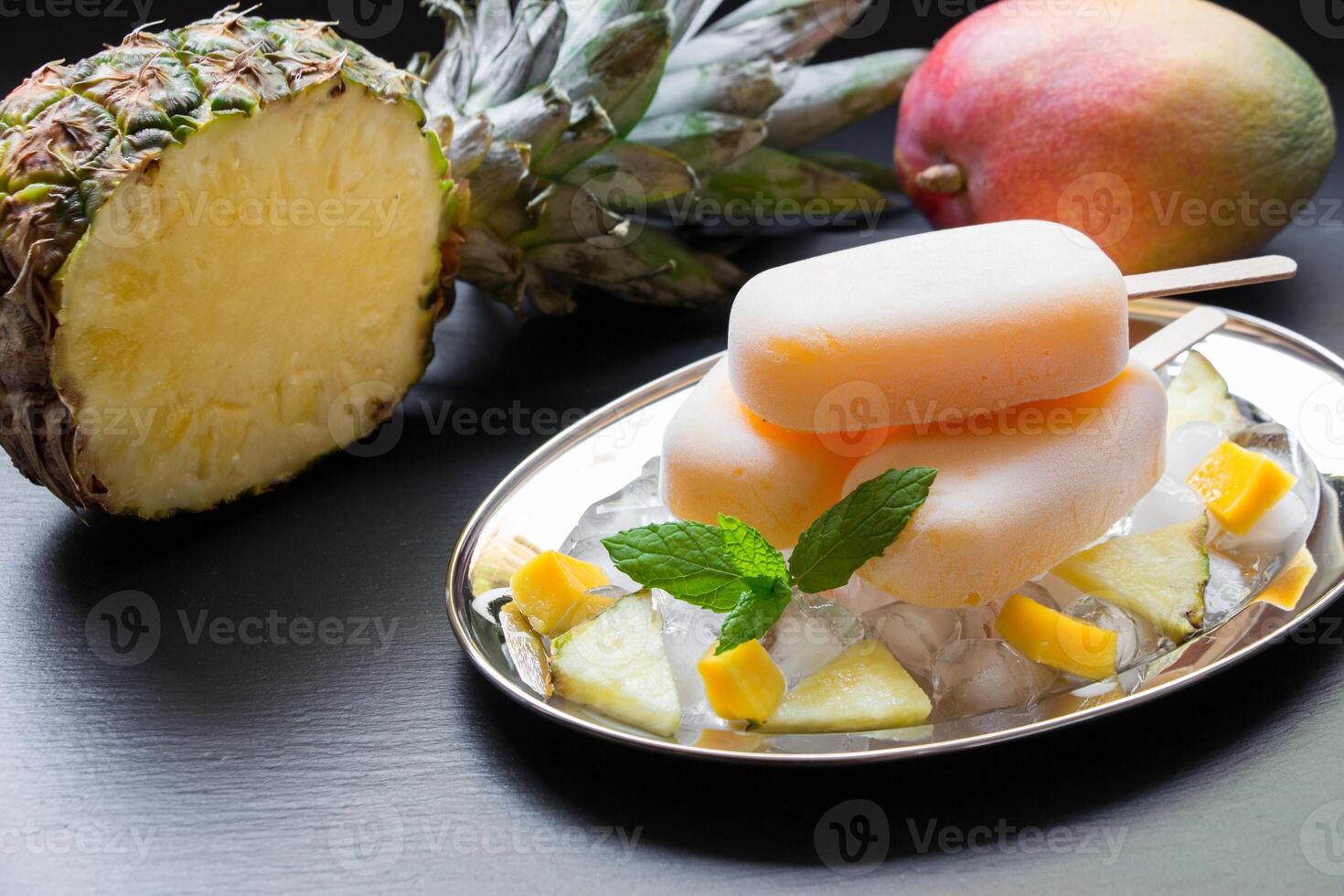 Exotic sorbet ice cream popsicles on silver plate with ice cubes chopped pineapple mango pieces, mint near half of pineapple, mango on a black background. Pineapple mango sorbet ice cream popsicles. photo