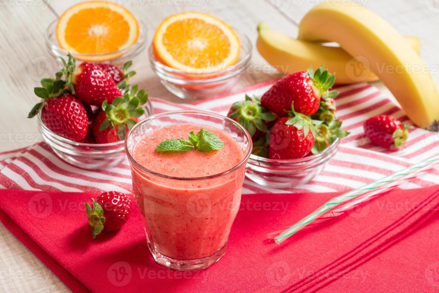 Fruit smoothie glass on red and white stripes napkins around spread ingredients. strawberries, sliced orange, bananas, blurred on white wood background. Horizontal. Daylight. photo