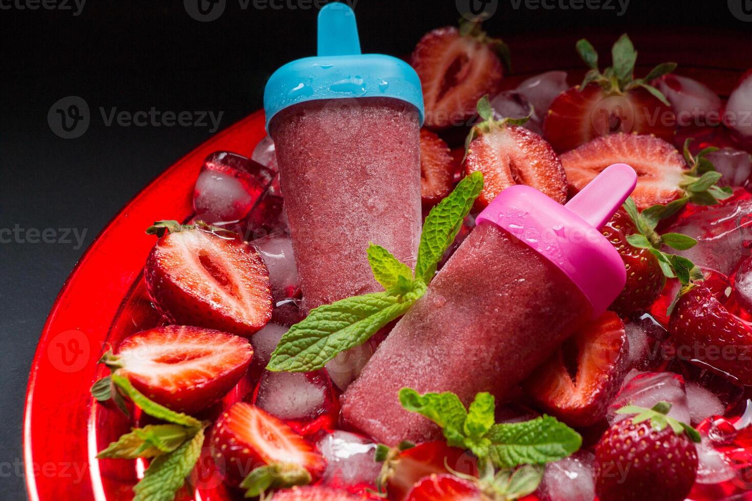 Two trawberry ice cream popsicles on red plate with ice cubes chopped strawberries and mint leaves on a black background. Strawberry ice cream popsicles. Horizontal. photo
