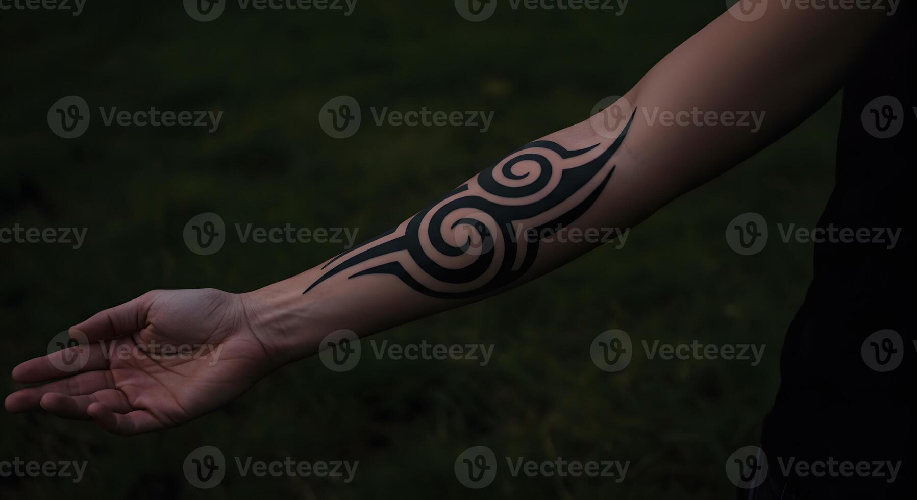 Black tribal tattoo on the forearm with swirling patterns shown against a dark grassy background during twilight photo