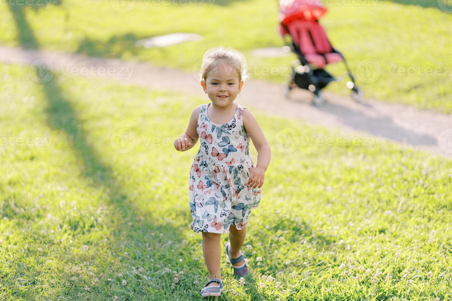 Little girl walks smiling along the green lawn from the stroller on the path photo