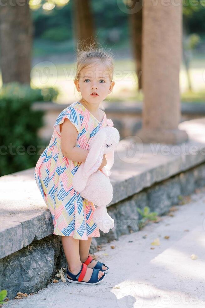 Little girl with a plush hare in her hands gets up from a bench in the park, turning her head to the side photo