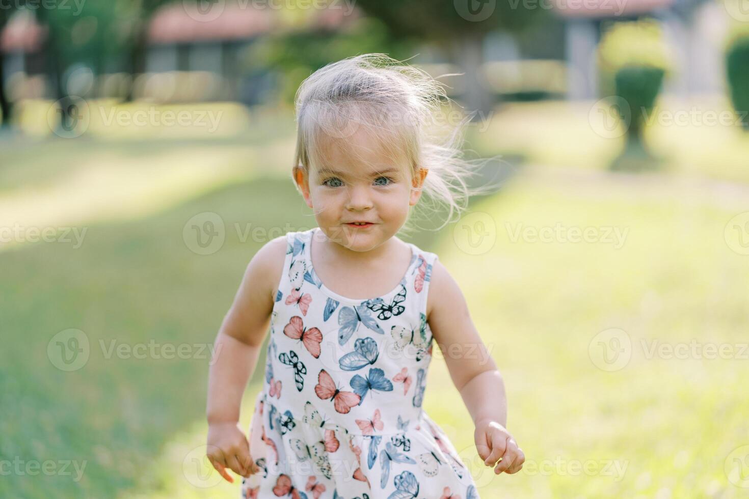 Little smiling girl with her hair fluttering in the wind stands on a green meadow photo