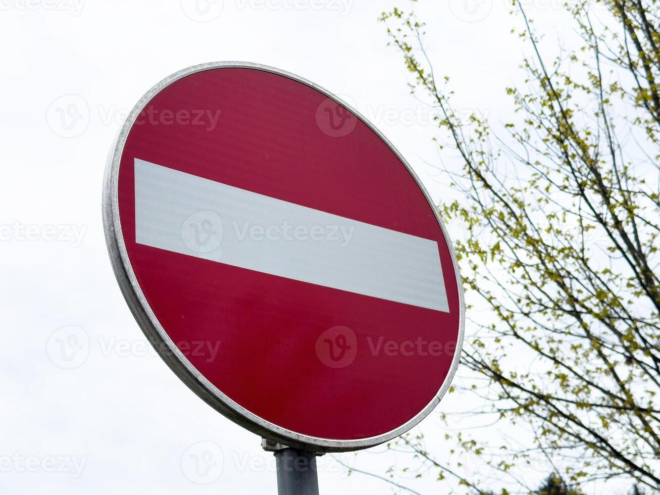Red do not enter sign against cloudy sky and leafless tree in background photo