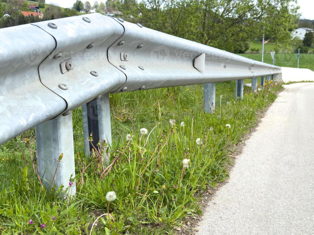 Close-up of roadside guardrail and dandelions along asphalt road in rural area photo