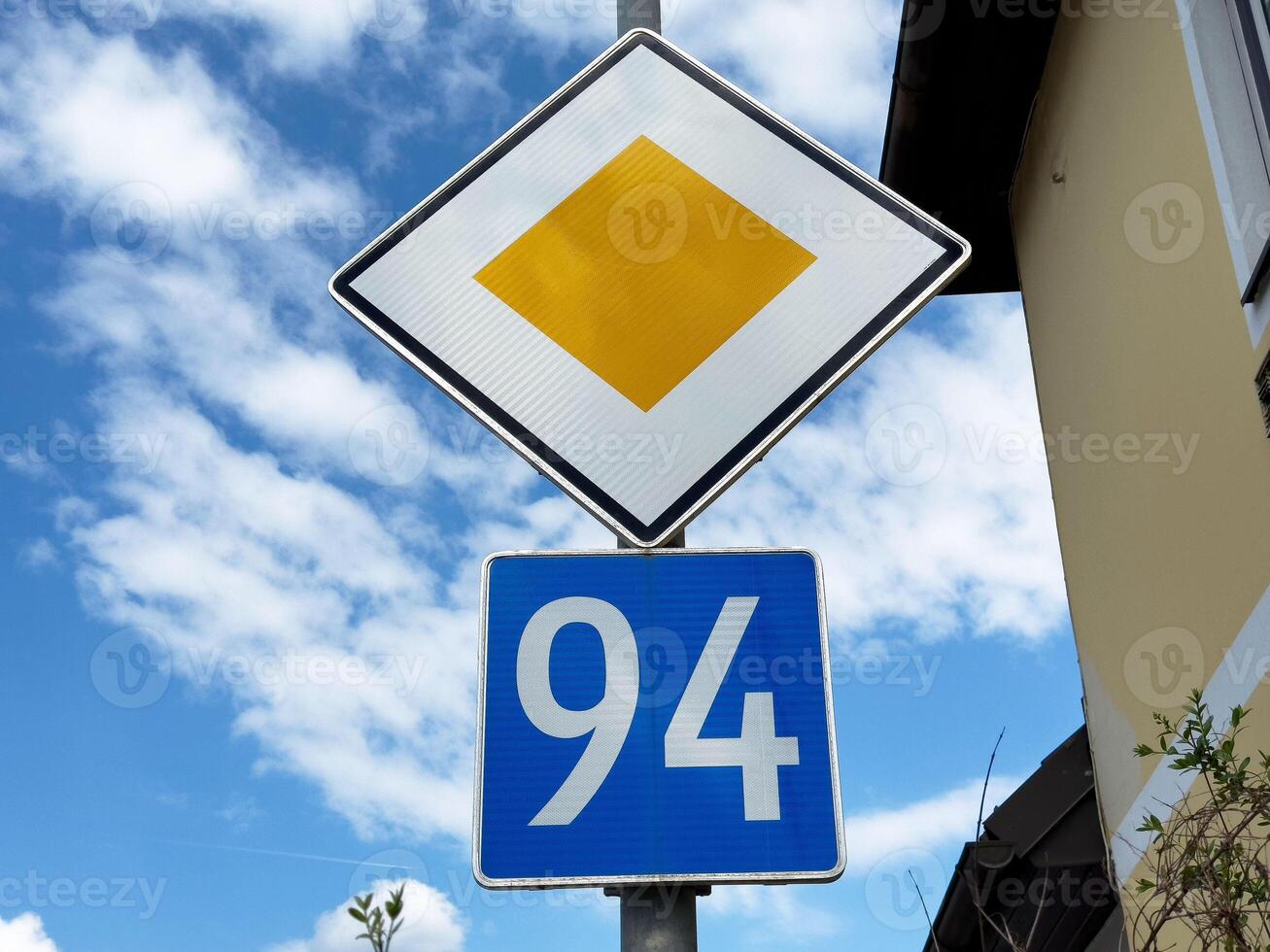 Yellow priority road sign with blue number 94 overhead on bright sky background photo