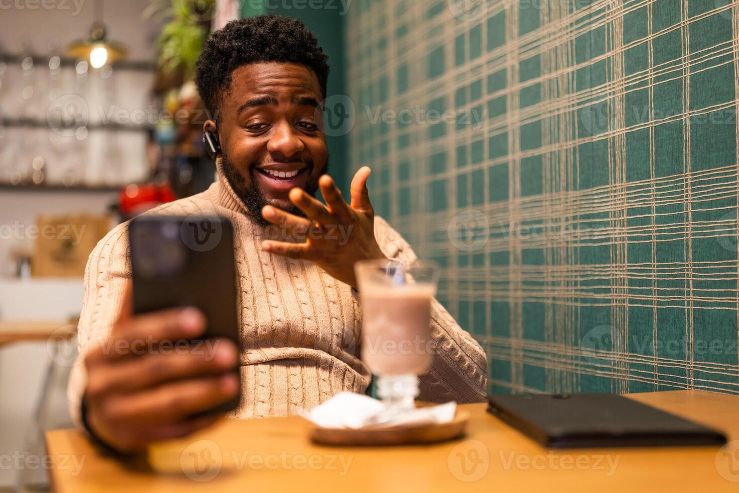 Cheerful black man in his 20s using a mobile phone with earphones, chatting online while relaxing at a cafe after work. photo