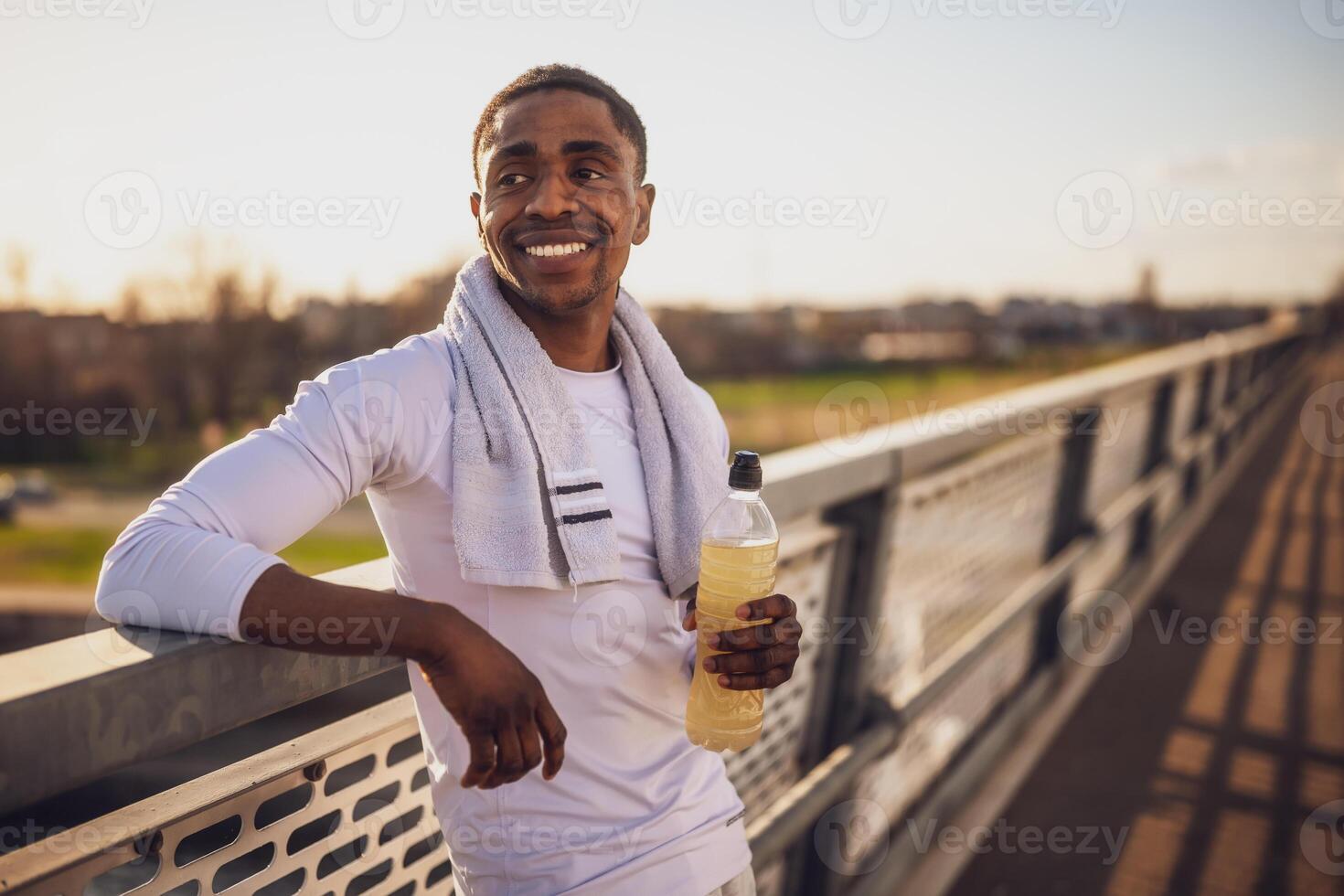 Muscular and fit, an African American man smiles while taking a break from his intense workout in the park, sipping juice to stay hydrated under the sunny sky. photo