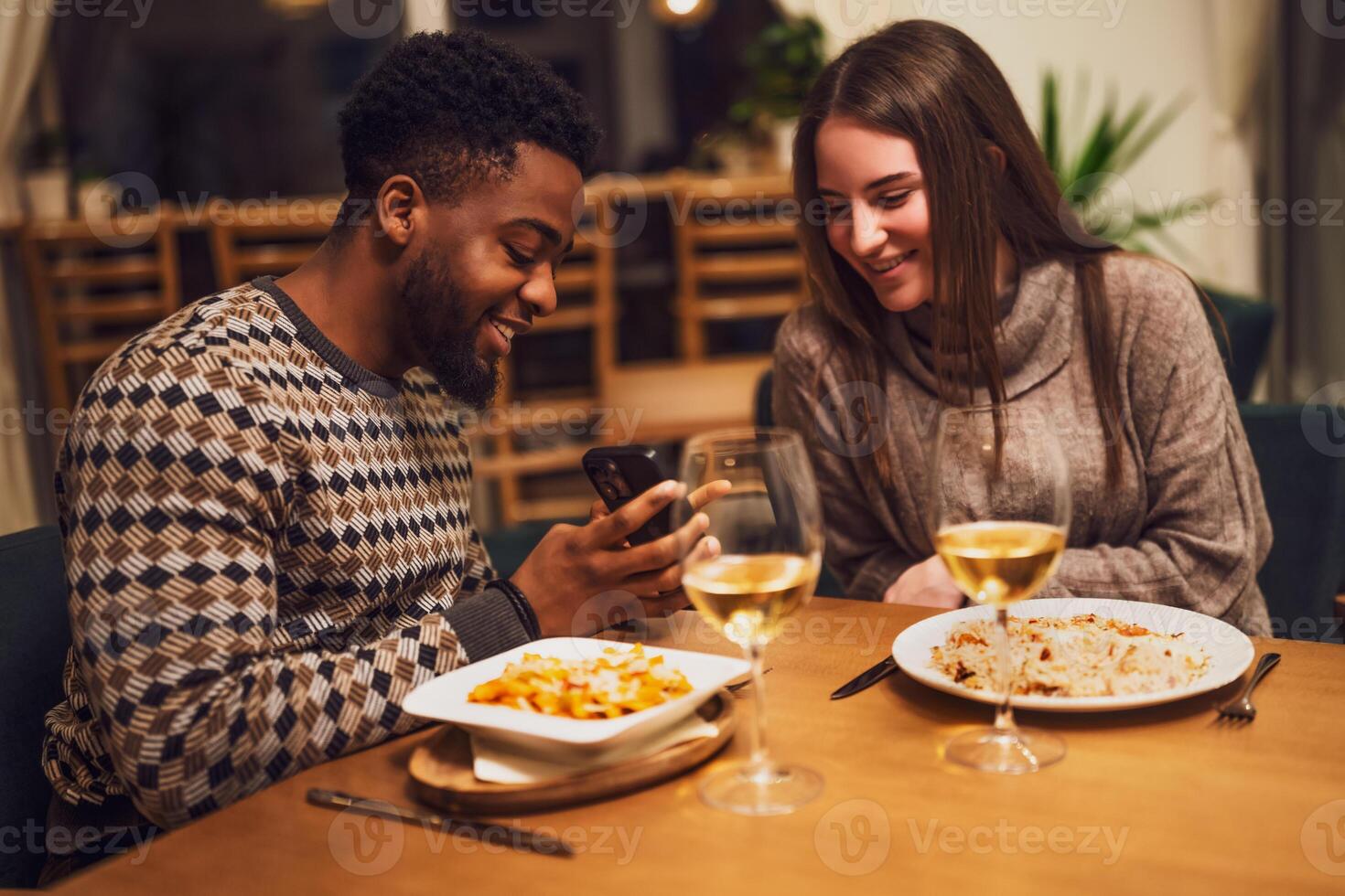 Happy friends sitting at table, smiling and talking while checking something on a smartphone during relaxed lunch in cozy restaurant. photo