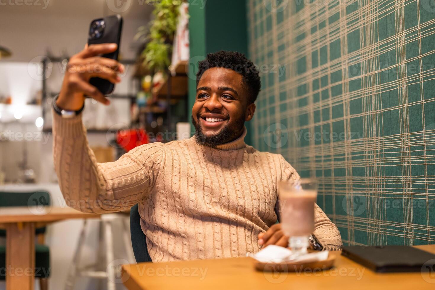 Portrait of a happy African man using his phone for a telecommuting call, sipping hot chocolate at a trendy bar. photo