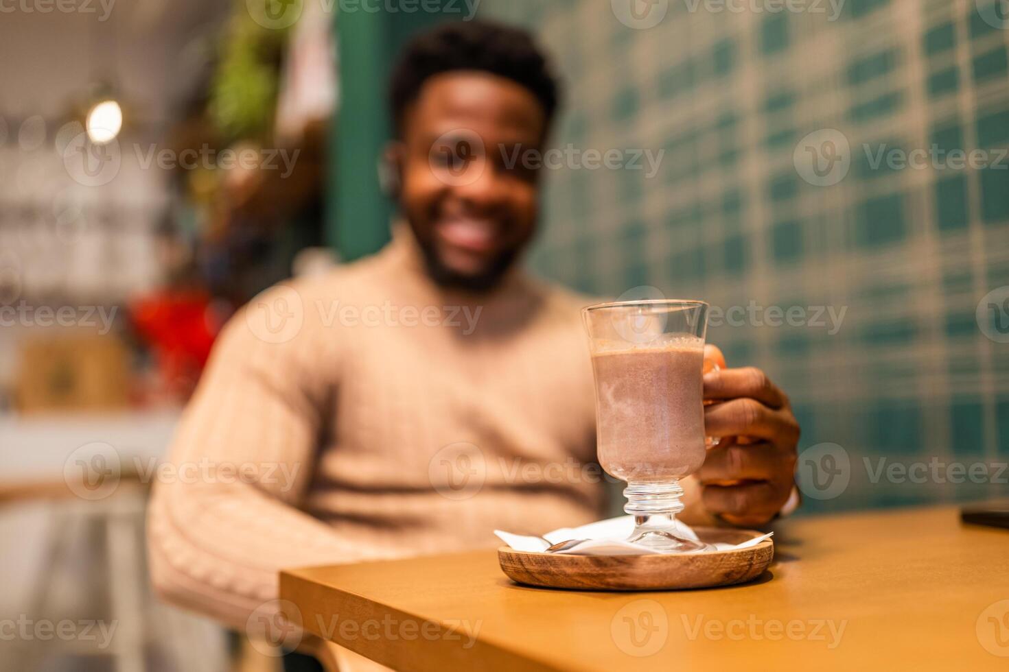 Young African man enjoying peaceful moment alone at cafe table, holding hot chocolate with relaxed smile. Selective focus on glass with hot chocolate. photo