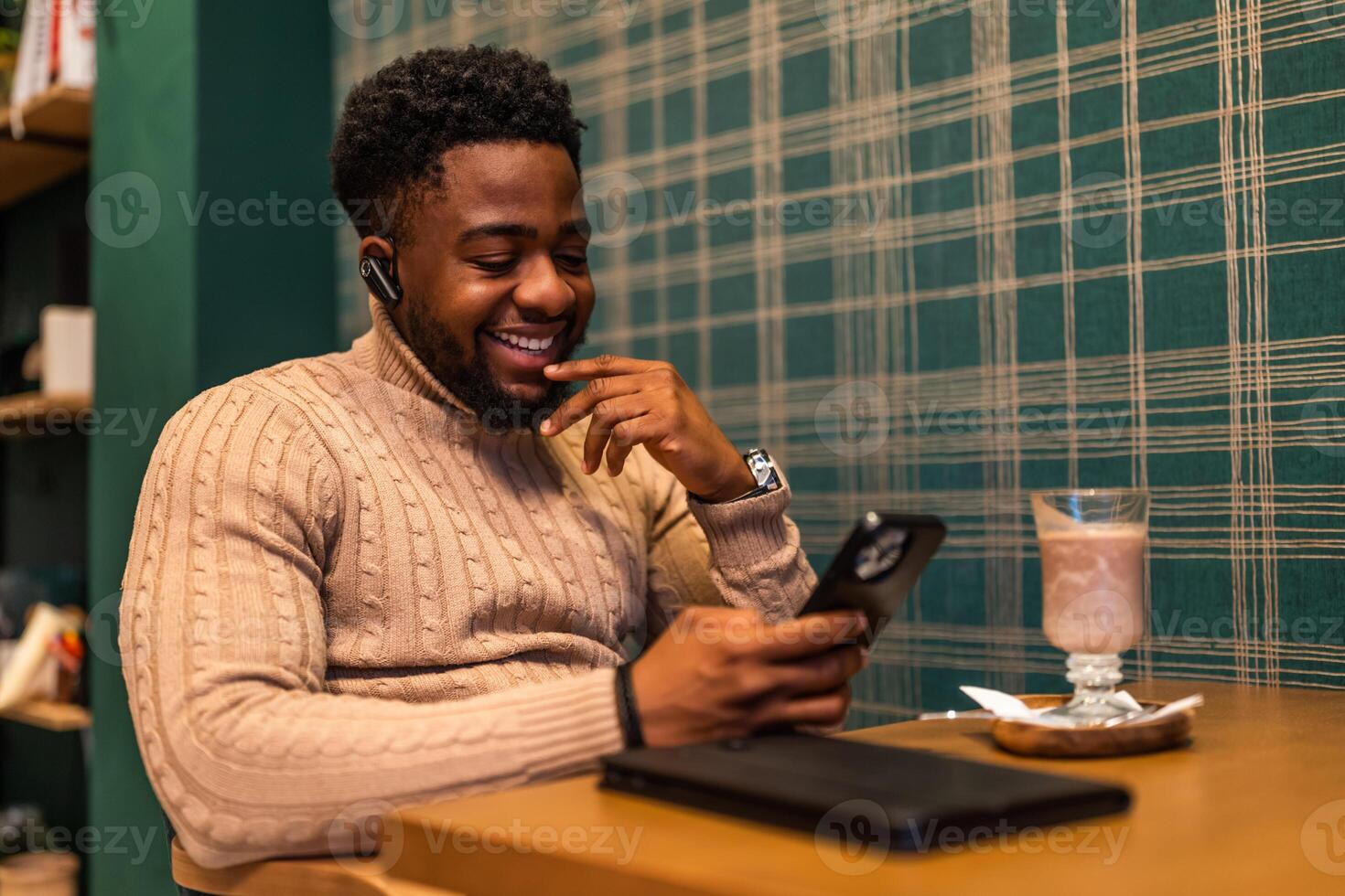 Smiling African freelancer sitting comfortably at cafe and enjoying a break while messaging on his smartphone. photo
