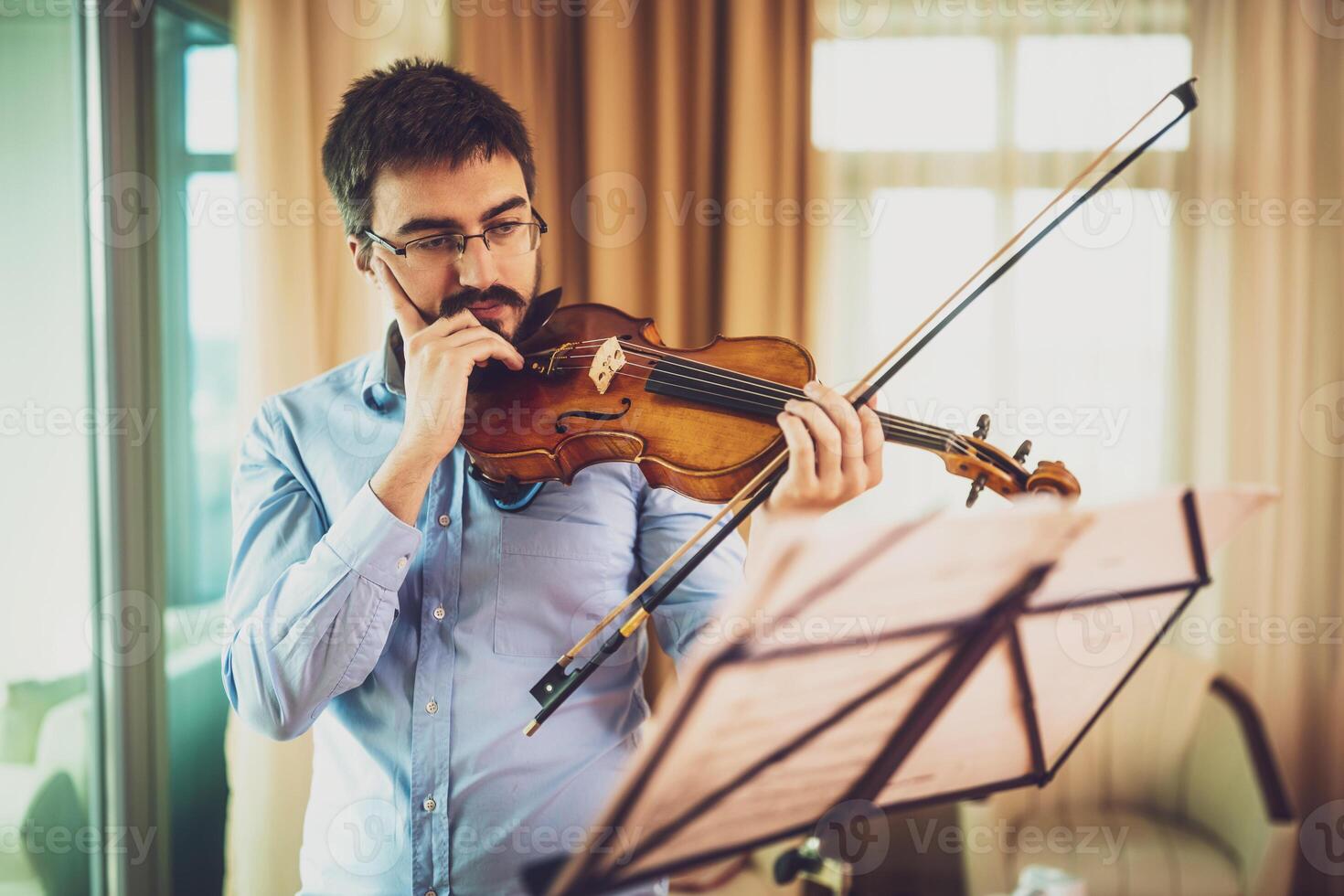Young male violinist practicing at home with intense focus, wearing eyeglasses and holding violin in quiet room. photo