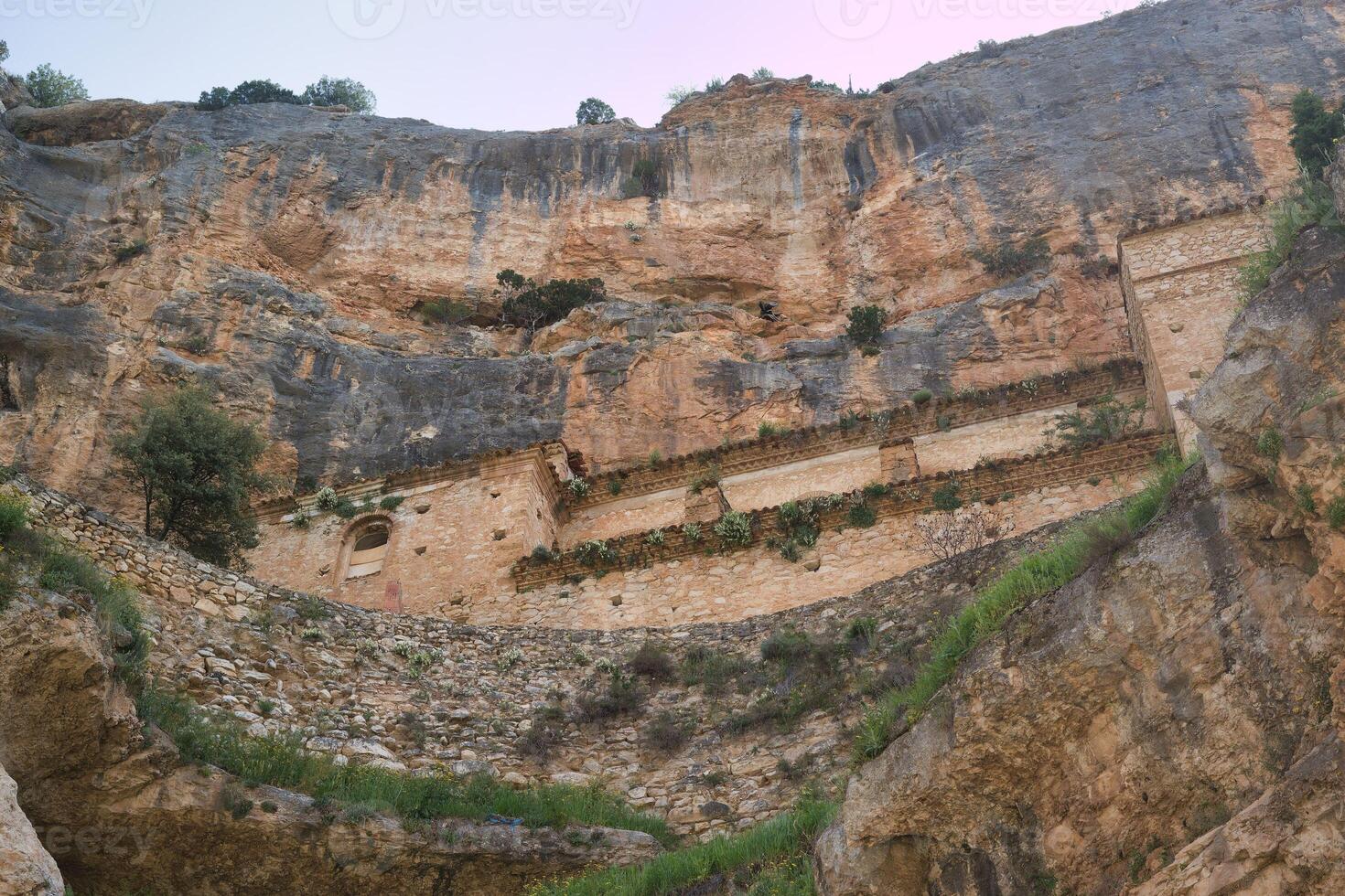 Ancient stone structures built into rocky cliffs overlooking a lush green valley in Spain at dusk photo