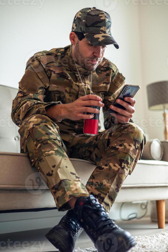 Depressed soldier wearing camouflage uniform sitting on sofa using smartphone and drinking energy drink photo