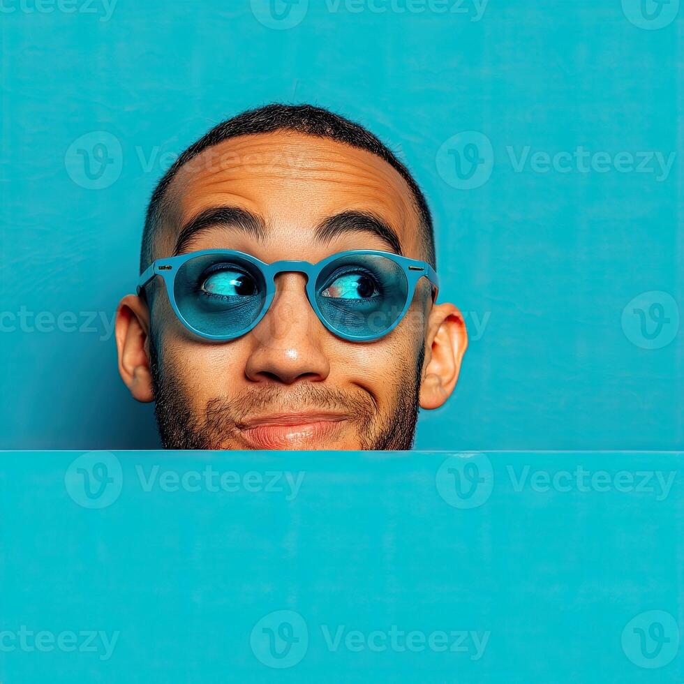 Curious man wearing blue sunglasses peeking over blue background photo