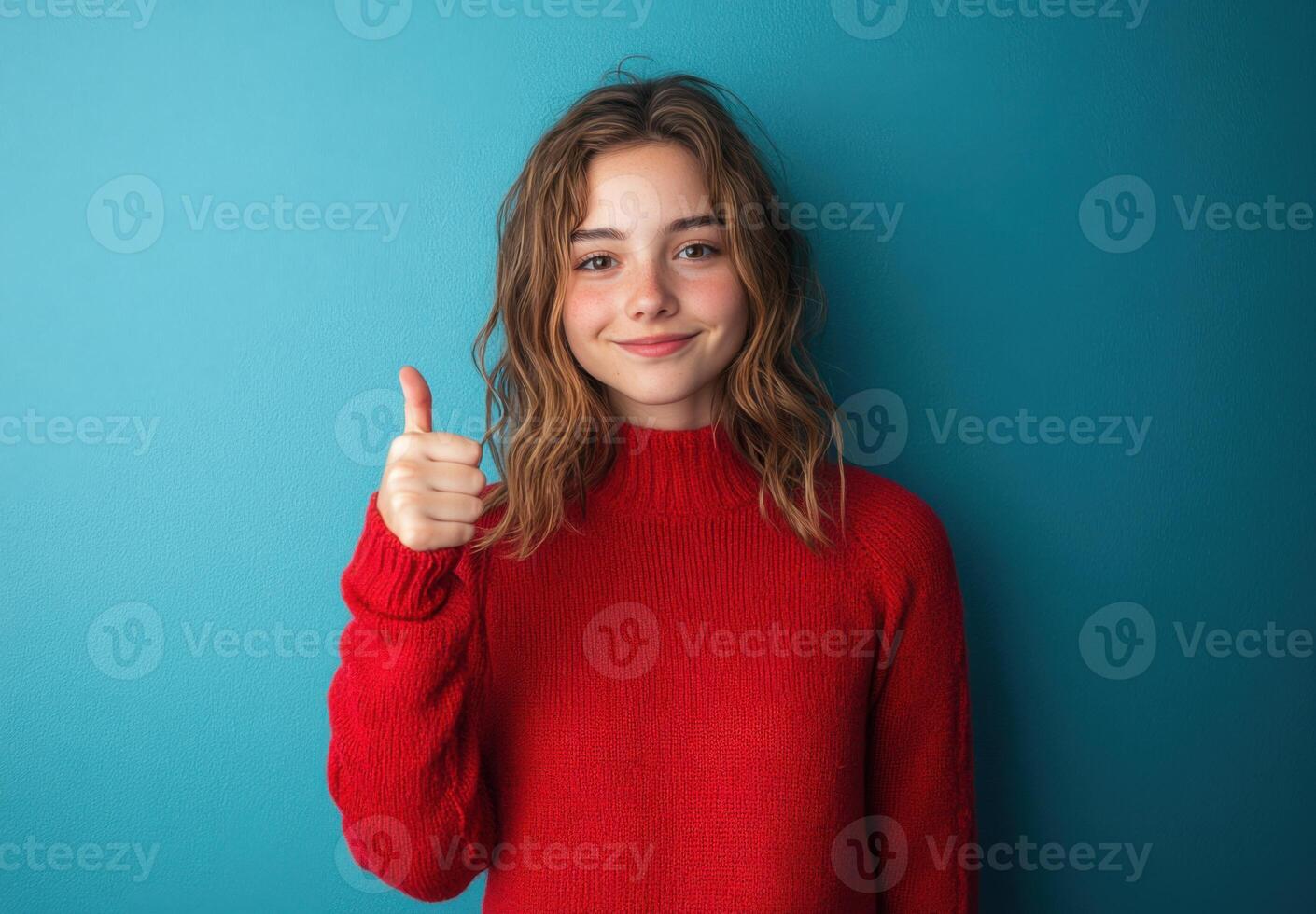 Portrait of a Smiling Teen Girl in Red Sweater Giving Thumbs Up Against a Teal Background photo