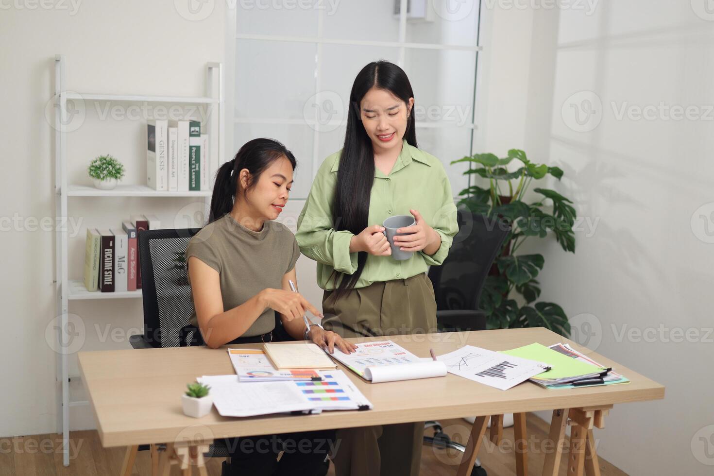 High angle view shot, Two female colleagues are working together in an office. A young woman is standing holding a cup of coffee and exchanging ideas with another colleague who is sitting. photo