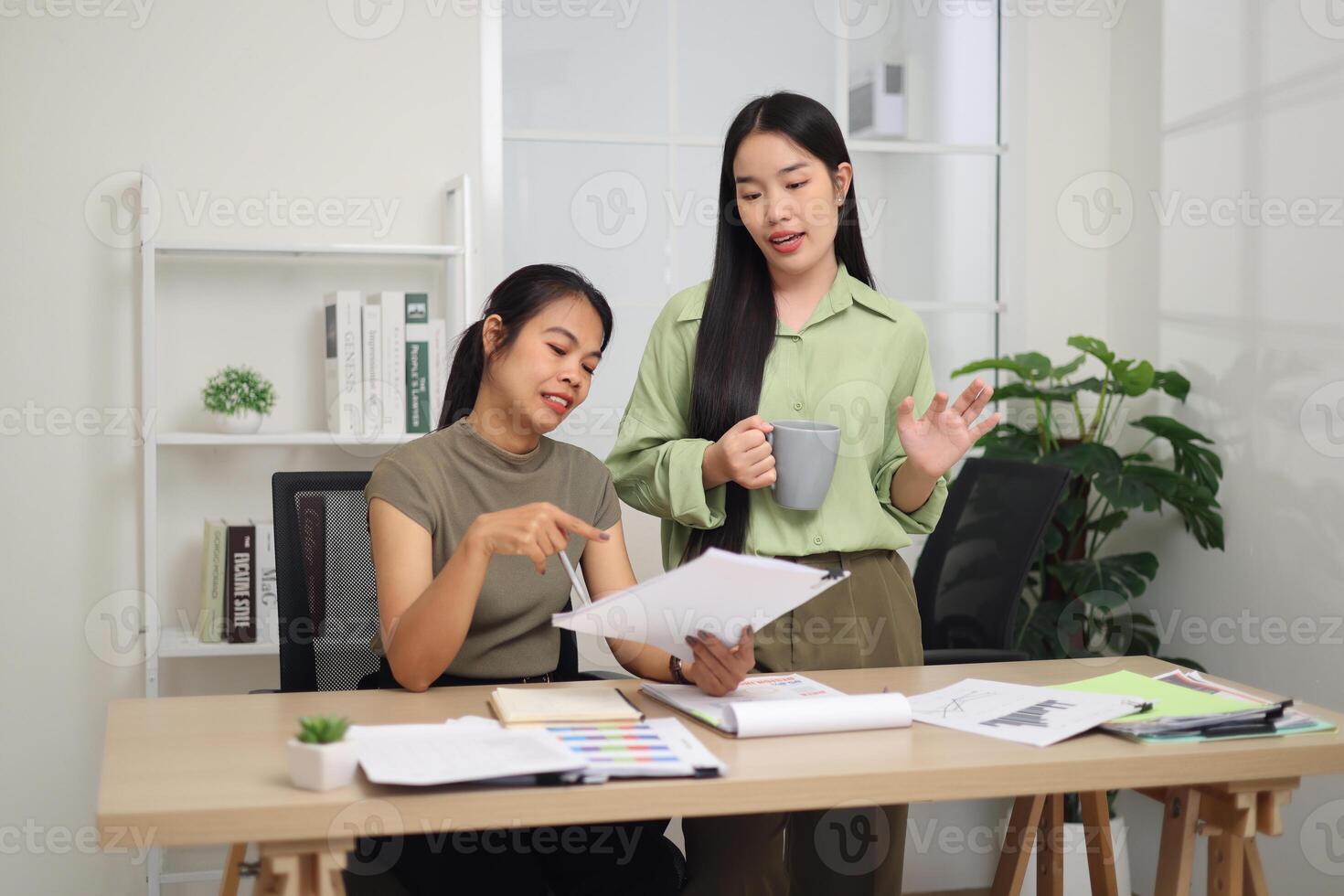 Two colleagues are working together in an office. A young woman is standing holding a cup of coffee and exchanging ideas with another colleague who is sitting. photo