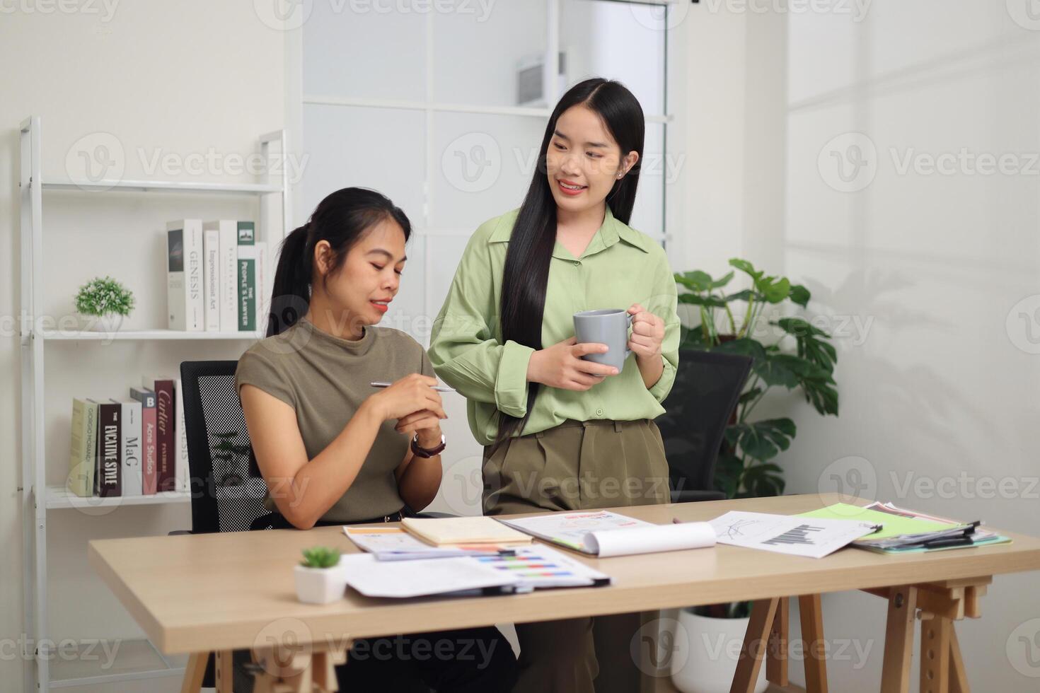 Portrait of Two colleagues are working together in an office. A young woman is standing holding a cup of coffee and exchanging ideas with another colleague who is sitting. photo
