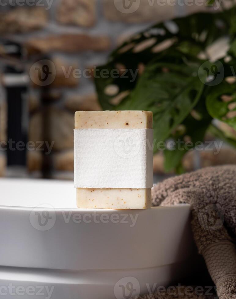 Soap bar with a blank label on vessel sink near grey towels and green plant close up, mockup photo