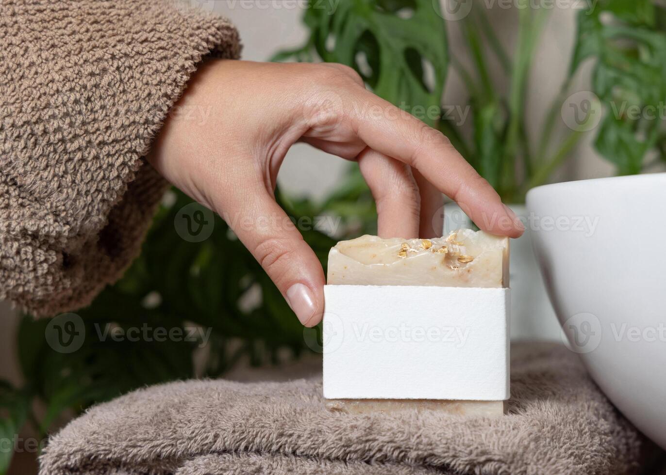 Closeup of female hand in brown bathrobe holding soap bar with blank label, mockup photo