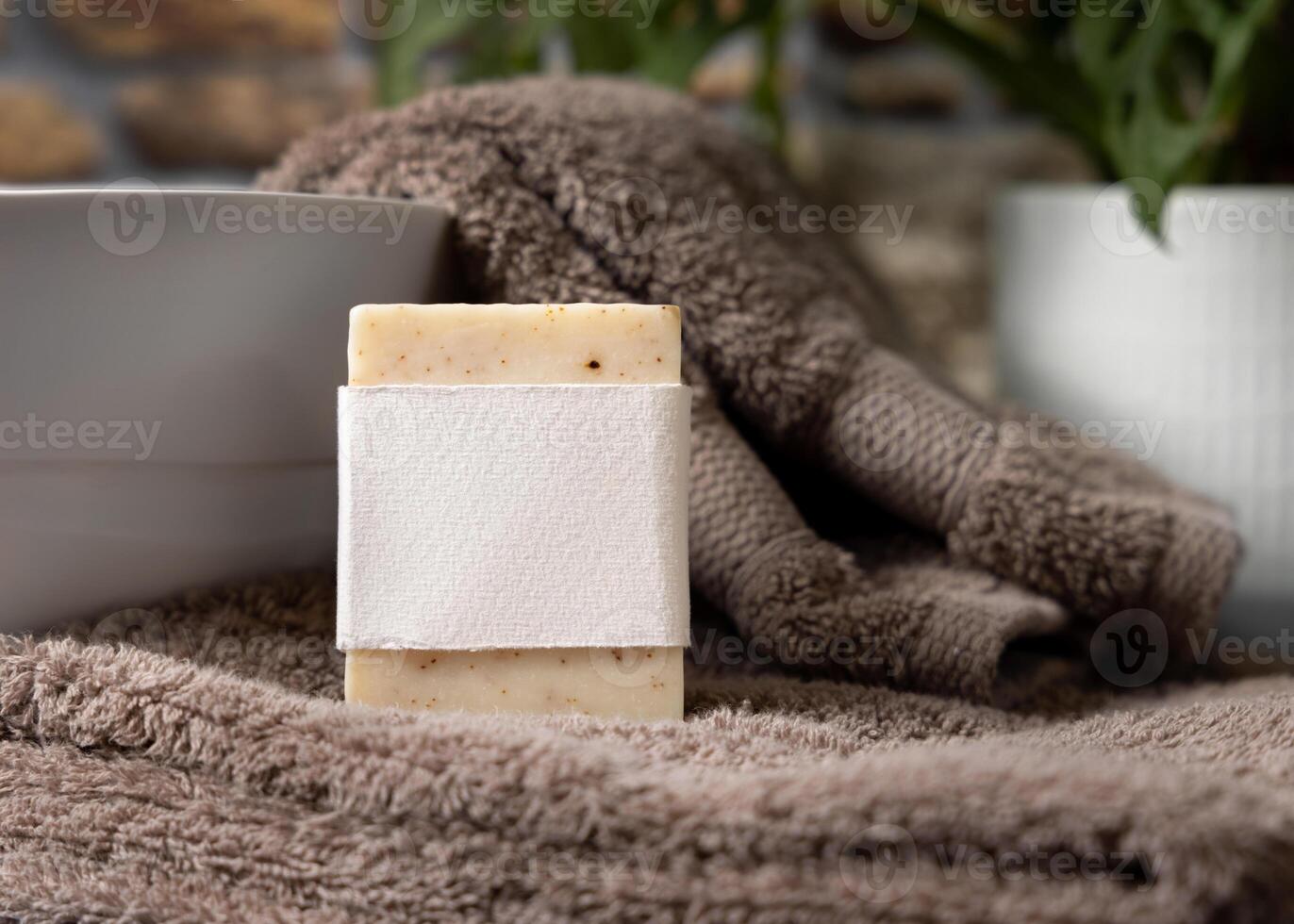 Soap bar with a label on brown folded towels near plant and basin in bathroom, mockup photo