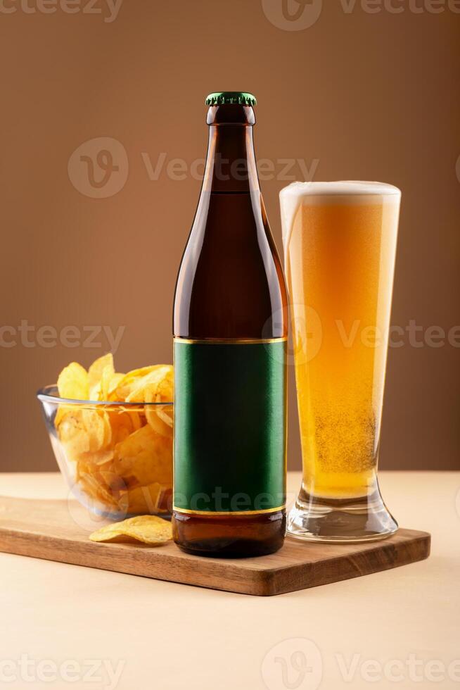 Beer bottle with green label, full beer glass and bowl of potato chips on wooden tray over beige background. Concept of bar ads, party visuals and beer snack combination promotions. Beer with chips photo